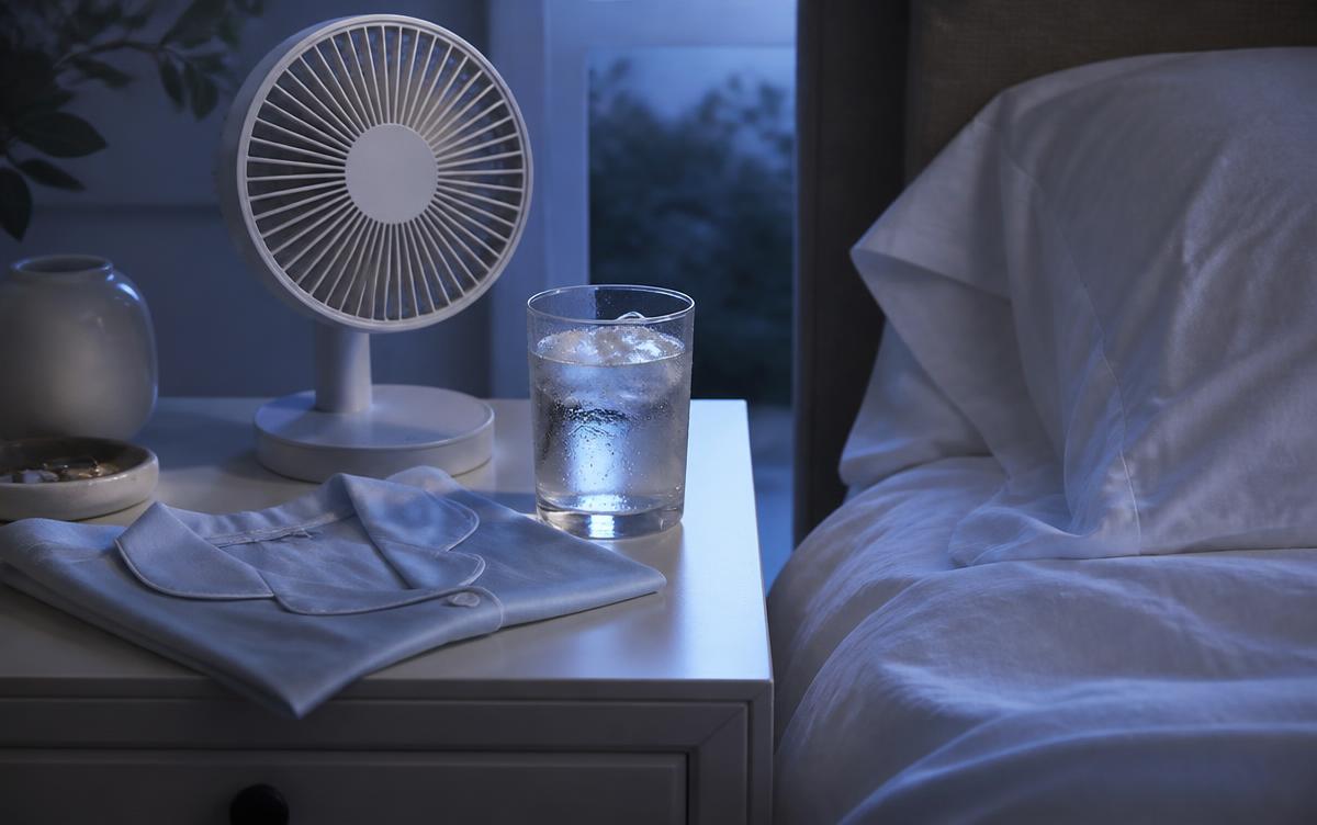 Mesa de cabeceira com ventilador pequeno, copo de água gelada e pijama de algodão dobrado ao lado de cama com roupa de cama branca em luz azulada de madrugada